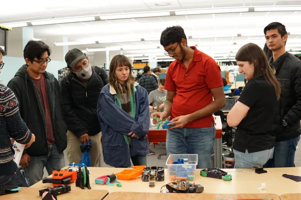 A group examines electronic components and 3D-printed parts on a table, with one person in red demonstrating.