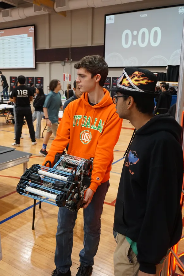 Two people are at a robotics competition, one holding a robot, while the other watches an ongoing match.