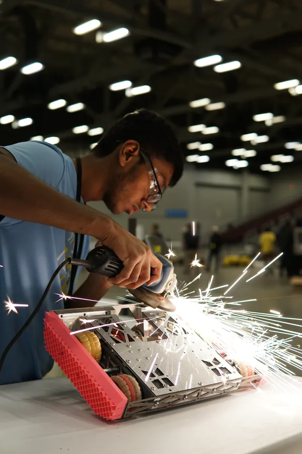 A person wearing safety glasses is using a power tool to cut a part on a robot, producing a shower of sparks.