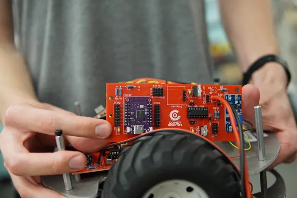 A student holds a custom-designed red printed circuit board with a white Comet Robotics logo printed at the center.