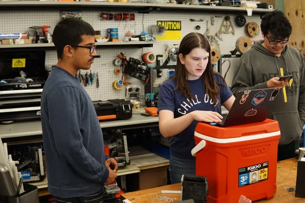 Two people work together, standing around a laptop on top of a red cooler.